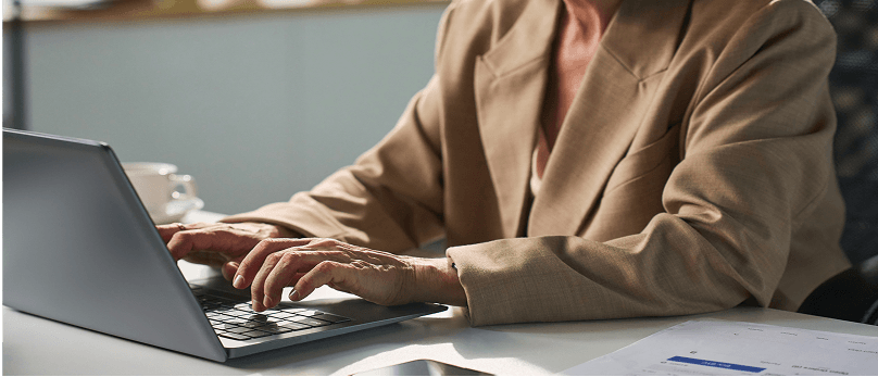 Eine Person, die einen beigen Blazer trägt und an einem Schreibtisch auf einem Laptop tippt.